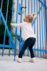 Little girl pulls the closed metal gates of the house towards herself. Side view
