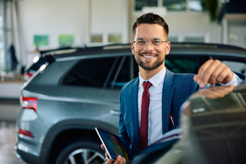 Happy salesman working at car dealership and looking at camera.