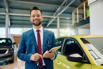 Happy car salesman with digital tablet in showroom looking at camera.