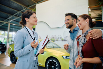 Happy saleswoman talks to young couple who is buying new car in showroom.