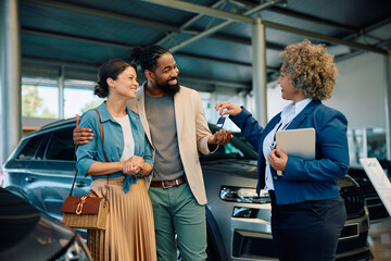 Happy multiracial couple receiving keys of their new car from saleswoman in showroom. - obrazy, fototapety, plakaty