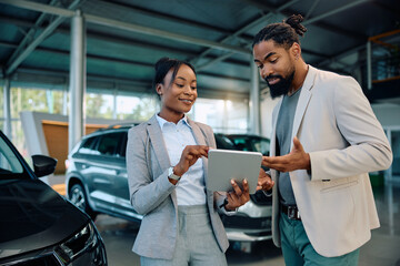 Black man and car saleswoman woman using digital tablet in showroom. - obrazy, fototapety, plakaty