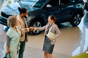 Happy saleswoman shaking hands with couple who is buying new car in showroom.