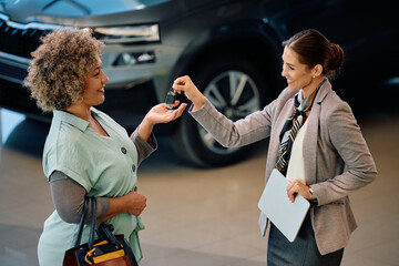 Happy black woman receiving keys from saleswoman after buying new car in showroom.