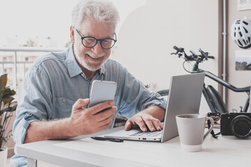 Smiling senior man with eyeglasses using phone and laptop together. Bearded white haired elderly man enjoying tech and social