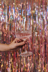 Persons hand holding glass of champagne wine over sparkling tinsel curtain. Christmas, New Year...