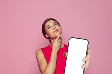 Naklejka premium Pretty woman thinking and holding smartphone with empty white screen display for new Application on pink studio wall background. Mockup banner