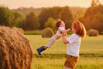 Mother holding her child, standing on a field with hay stacks amidst the setting sun, while the...