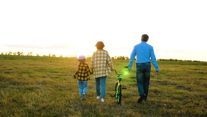 Mom and dad guide little child through sunlit field carrying colorful bicycle