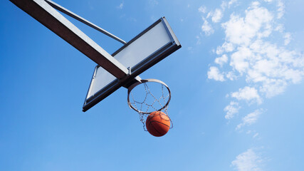 A basketball backboard seen from the back and below, low angle shot looking up at basketball going through basketball hoop, cloudy blue sky background