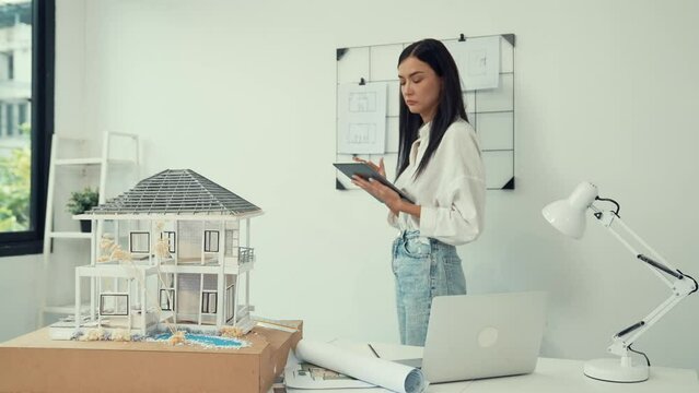 Beautiful caucasian architect focus on using tablet to check house model on meeting table with blueprint, house model and architectural document scatter around at meeting room. Immaculate.