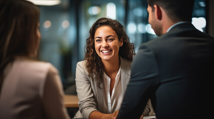 Smiling woman in a business meeting, wearing a blazer and a white shirt, engaged in a conversation with colleagues who are partially visible and out of focus in the foreground. - obrazy, fototapety, plakaty