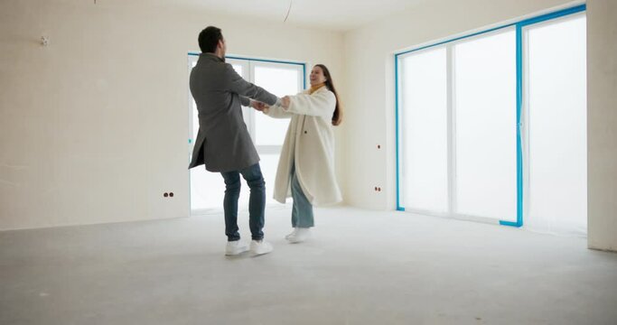 The couple hold hands and spin around their axis in circles. Joy, happiness of cash purchase of house from developer, empty middle of apartment, shell of apartment.