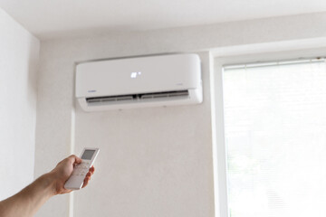 Hand holding remote control aimed at the air conditioner. Young man turning on air conditioner with...