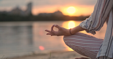 Woman practicing yoga, joins her index finger and thumb to sign apana mudra. Sunset, river, beach, orange sun reflected on surface water.
