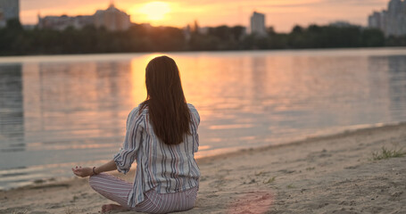Girl doing yoga sitting on sandy beach river. Lotus pose, join palms hands above head. Back view, sunset rays sun.