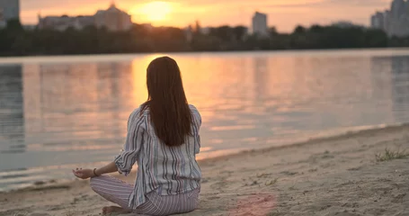 Rucksack Girl doing yoga sitting on sandy beach river. Lotus pose, join palms hands above head. Back view, sunset rays sun. © Oleksii