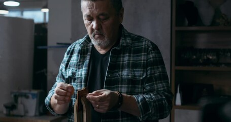 successful mature craftsman restores items made from leather and leather material. bearded Asian man concentrated on sewing at workshop.serious professional craftsman takes needle run through hole - Powered by Adobe