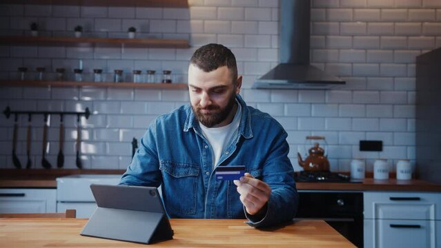 Young adult man sitting against the kitchen counter entering credit card number on tablet device for makes secure easy distant electronic payment at home