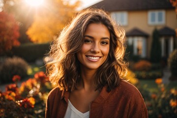 Obraz premium Smiling young brunette posing at a beautiful garden looking at the camera during late autumn sunset with a sun flares in the background, surrounded by friends and relatives.
