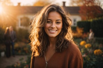 Obraz premium Smiling young brunette posing at a beautiful garden looking at the camera during late autumn sunset with a sun flares in the background, surrounded by friends and relatives.