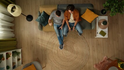 Top view shot of a homosexual couple at home. They are sitting on the couch, playing videogame with gamepads in their hands, having fun and play fighting.