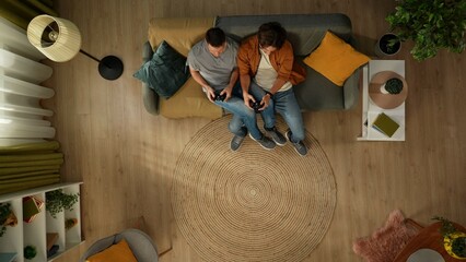 Top view shot of a homosexual couple at home. They are sitting on the couch, playing videogame with gamepads in their hands, having fun and play fighting.