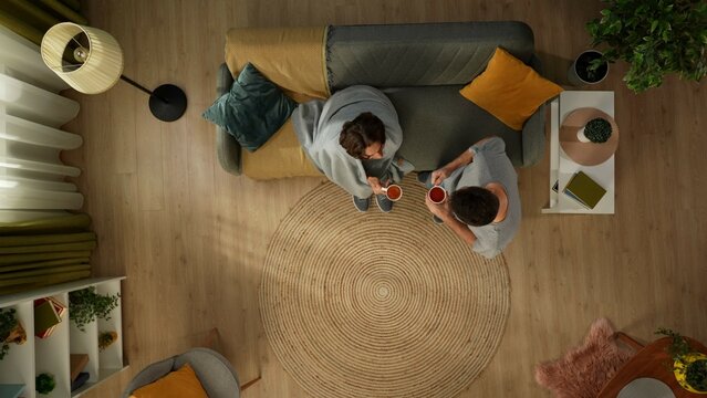 Top view shot of a homosexual couple at home. One is sitting on the couch, wrapped in a blanket. His partner is bringing two cups of tea to warm him up.