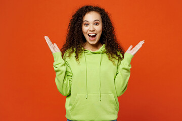 Young surprised shocked amazed overjoyed woman of African American ethnicity she wears green hoody casual clothes look camera spread hands isolated on plain red orange background. Lifestyle concept.