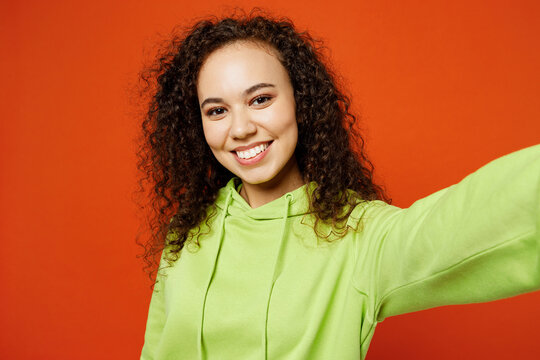 Close up young woman of African American ethnicity she wear green hoody casual clothes doing selfie shot pov on mobile cell phone look camera isolated on plain red orange background Lifestyle concept