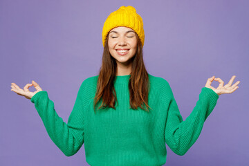 Young happy woman she wear green sweater yellow hat casual clothes hold spreading hands in yoga om aum gesture relax meditate try to calm down isolated on plain pastel light purple background studio.