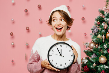 Merry smiling young woman wearing white sweater hat posing hold in hand clock look aside on area...