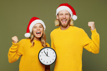 Merry young couple two friends man woman wears sweater Santa hat posing hold in hand clock do...