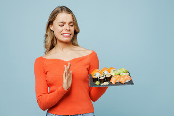 Young displeased sad woman wear orange casual clothes showing stop palm gesture holding eat raw fresh sushi roll served on black plate Japanese food isolated on plain blue background studio portrait.