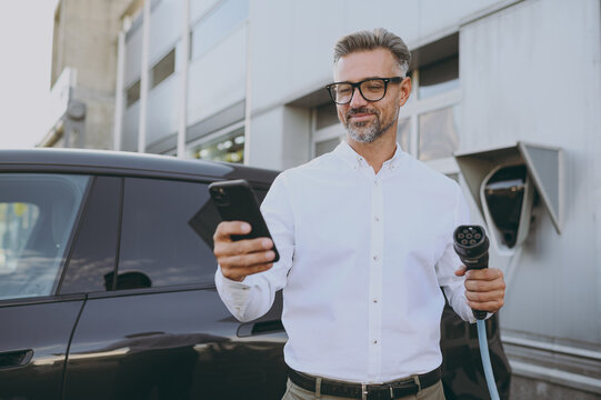 Adult confident man wearing shirt use mobile cell phone hold in hand electric car charger connect to electric car auto at charging station outdoor. Green energy solution for automobile fuel concept. - Powered by Adobe