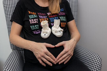 Kobieta w ciąży trzymające ręce w kształcie serca na brzuszku, na zdjęciu sa malutkie buciki. Koncept wychowywania i kosztów wychowywania dzieci.	
