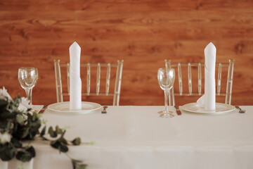 Table setting with a white tablecloth, white plates and clear glasses. The background is a wooden wall. A concept for creating a cozy and romantic atmosphere or a gourmet dinner.
