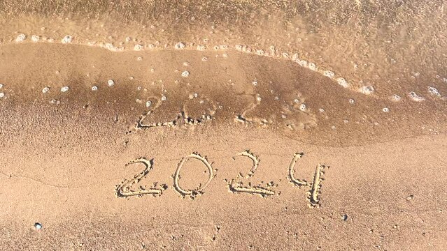 The concept of the New Year. The text of 2024, handwritten on a sandy surface. The figures of 2023 are washed away by the sea wave. Welcoming the year 2024. Happy New Year. Seashore, waves, sand.
