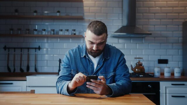 Bearded young adult man sitting against the kitchen counter holding a smartphone enjoying surfing internet, using social media or app