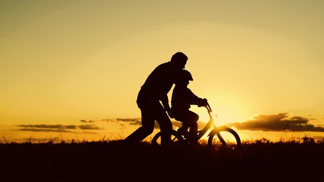 Father teaches child wearing safety helmet to ride bicycle Family in park. Child rides bicycle. Father helps his daughter ride bike. Kid, dad play together, sunset. Child dream learns to ride bicycle