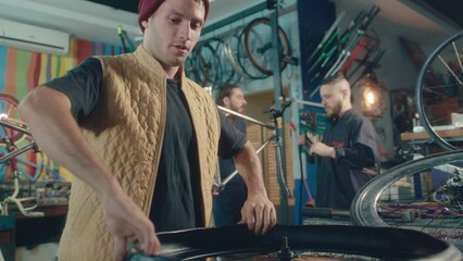 Young male mechanic changing tire on bicycle wheel, chatting with colleague and smiling during workday in repair shop - Powered by Adobe