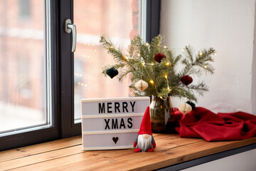 holidays, decoration and celebration concept - close up of merry christmas greeting on light box and gnome on window sill at home