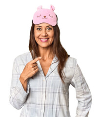 Middle-aged Caucasian woman in pajamas and mask in studio smiling and pointing aside, showing something at blank space.