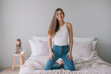 cheerful young girl with long hair in jeans sitting on the bed looking at the camera rejoicing in...