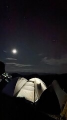Camping under the moonlight, with a tent pitched and a campfire glowing in the middle of the night. - obrazy, fototapety, plakaty