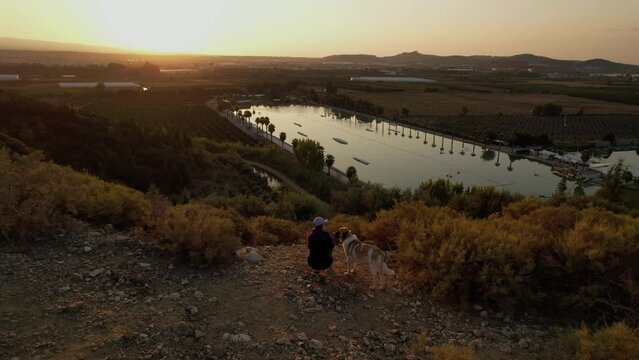 Girl with a dog are on top of a hill and watching the dawn. Against the background of a lake, wakepark, palm trees, plantations and greenhouses. Golden hour, sunrise. High quality 4k drone footage