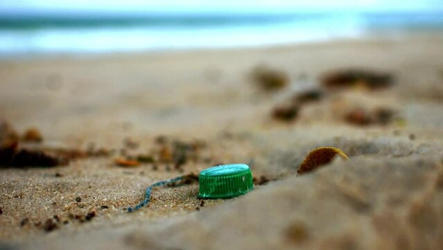 Green Plastic Bottle Cap in the Sand at a Windy Beach in Brazil with Waves Moving at the Back, Ocean Microplastics Problem, Sad Cold Colors