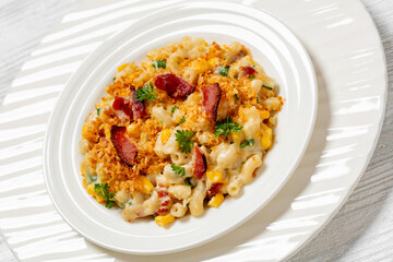 portion of mac and cheese with corn, bacon topped with panko breadcrumbs on white plate on white wooden table, dutch angle view, close-up