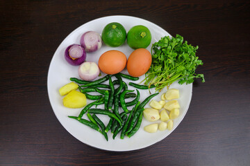 Eggs, lemons, onions, ginger, green chilies, coriander leaves and garlic are neatly arranged on a white plate on a black background.