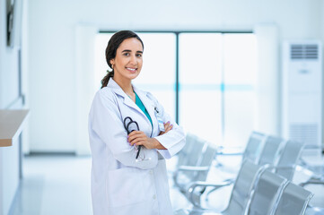 Attractive smiling female doctor wearing a white medical gown and holding a stethoscope, standing in the hospital corridor. Medicine and health care concept with copy space. looking at camera.